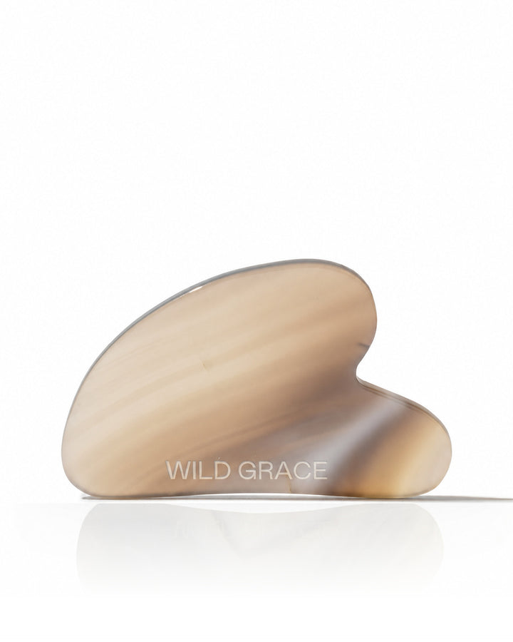 A curved, hand-shaped jade Gua sha massage tool with a translucent beige and light brown marbled pattern, resting on a reflective white surface against a white background. The brand name &