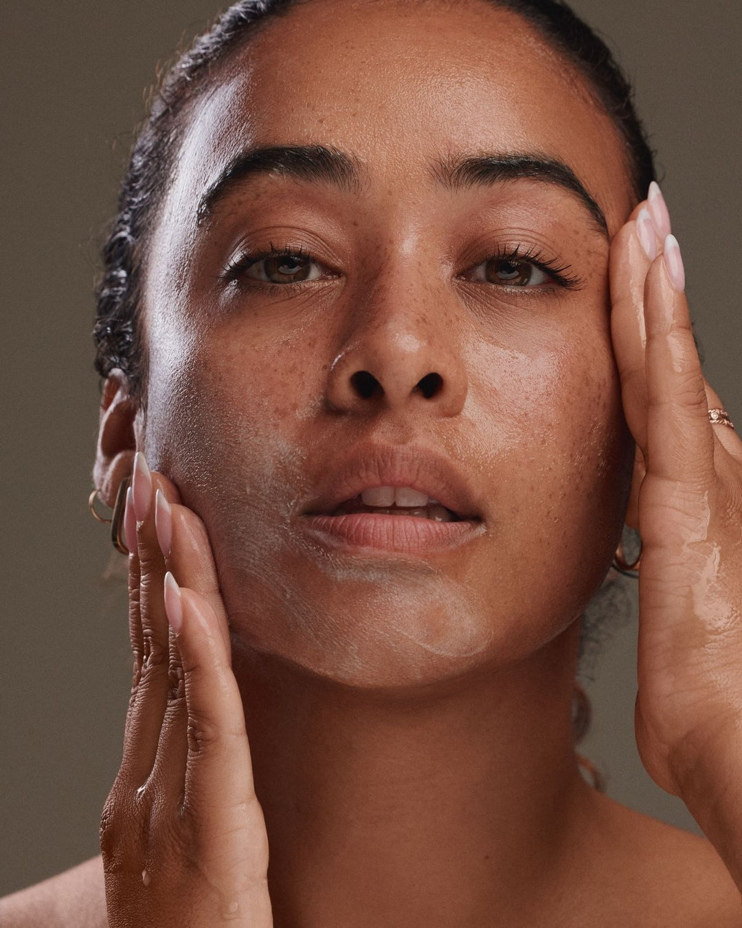 Close-up of a woman with her hands on her face, showing a white skincare cream or powder being applied. The background is a solid, warm taupe-brown color.