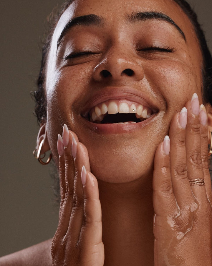 A close-up lifestyle shot of a woman with closed eyes and a wide smile, gently touching her face with both hands. She is wearing gold hoop earrings and a gold ring, and her skin appears to be glowing with a soft, radiant finish.