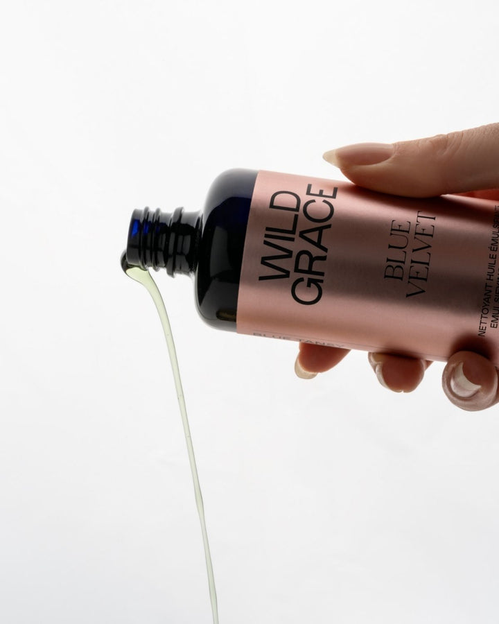 Hand holding a bottle of Wild Grace Blue Velvet emulsion with product being poured out.