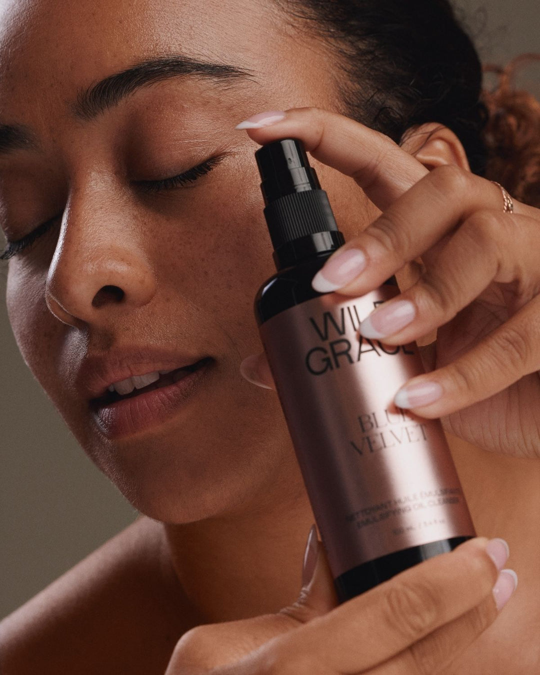 A close-up lifestyle shot of a woman holding a bottle of Wild Grace Black Velvet Hair Spray against her face, showing the product being used.