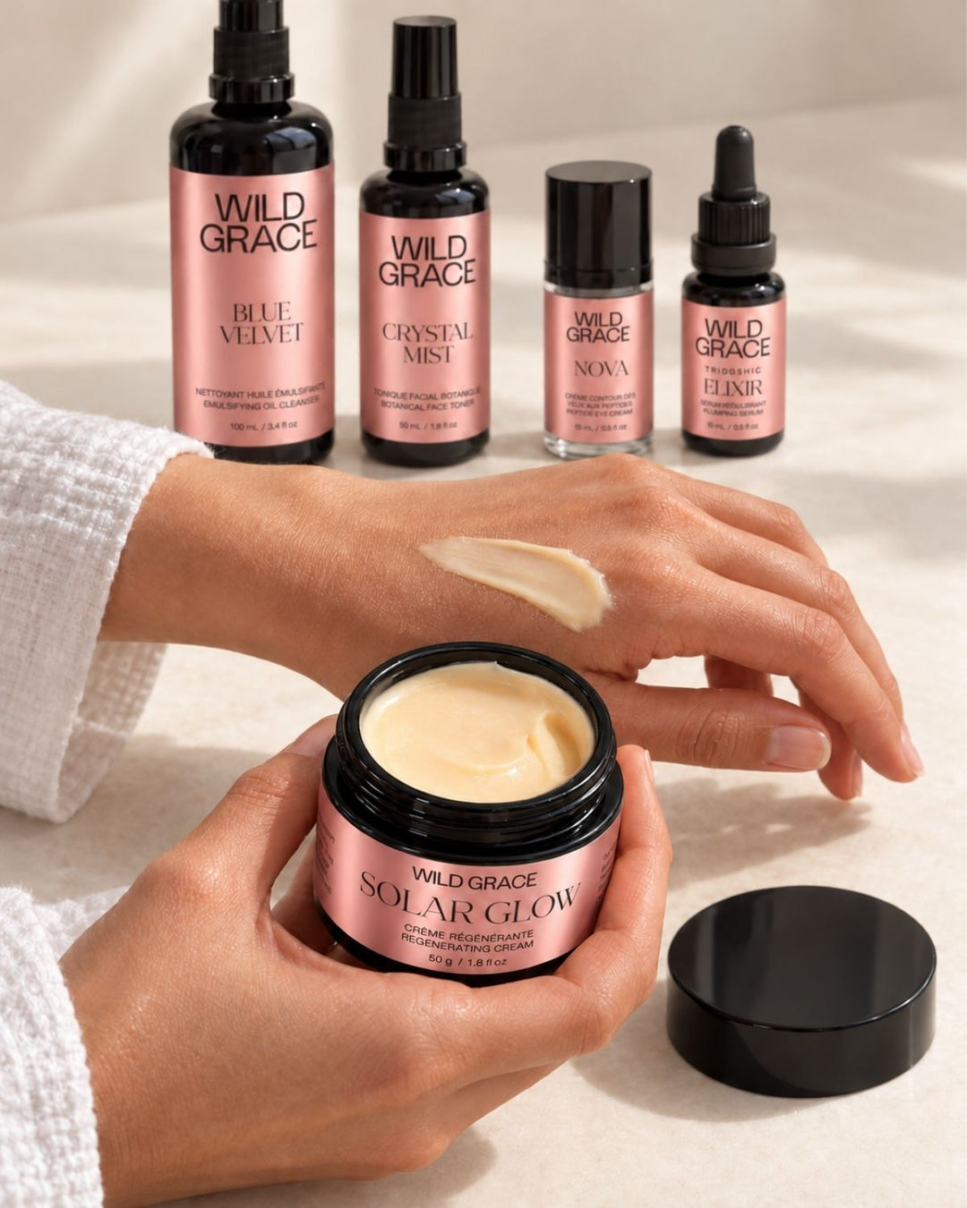 A person holds an open jar of Wild Grace Solar Glow face mask, showing the creamy beige product on their hand, with four Wild Grace skincare bottles lined up in the background on a light beige surface.