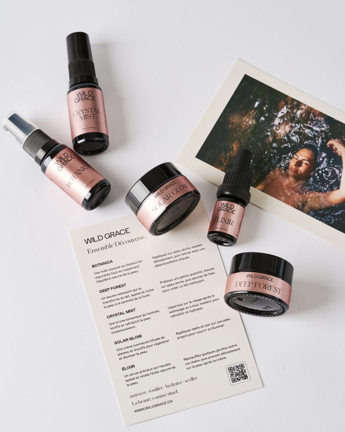 A flat-lay product arrangement on a white background featuring several Rose Crystal Infused essential oil bottles, a jar, a dropper bottle, a printed card, and a small printed photo. The products have rose gold labels with black caps and are branded 'Wild Grace'.