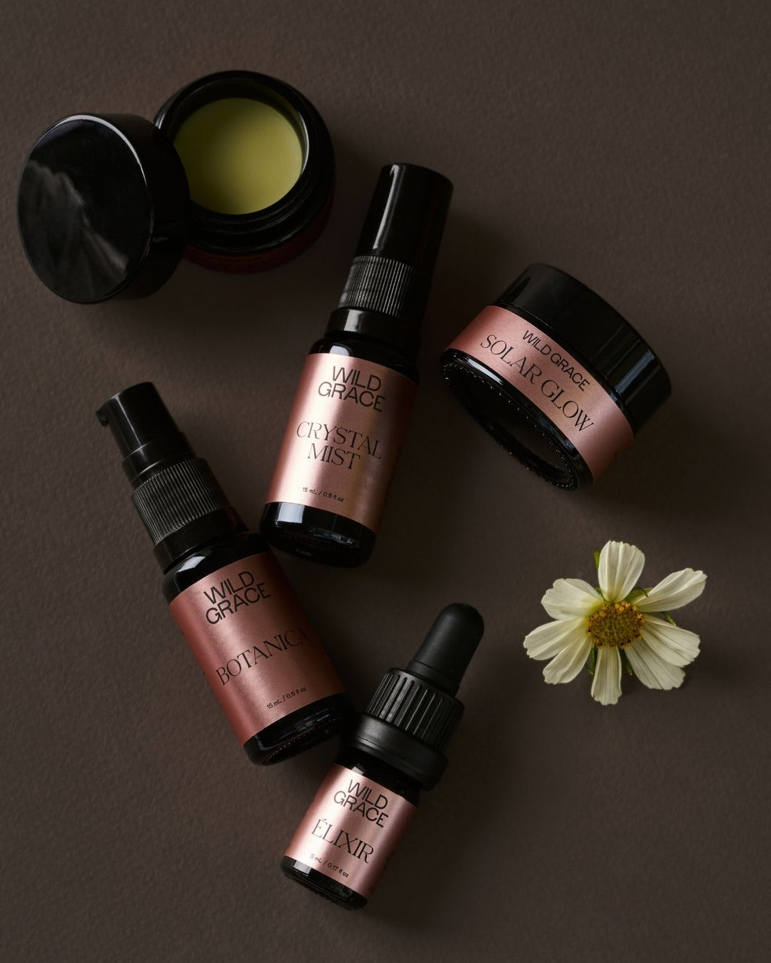 Four Wild Grace skincare products arranged on a dark brown background with a small white flower prop: an open jar of balm, a spray bottle labeled Crystal Mist, a dropper bottle labeled Hydrating, and a dropper bottle labeled Elixir. The products have black packaging with metallic rose-gold labels.