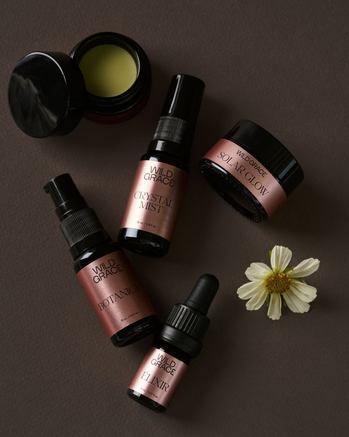 Four Wild Grace skincare products arranged on a dark brown background with a small white flower prop: an open jar of balm, a spray bottle labeled Crystal Mist, a dropper bottle labeled Hydrating, and a dropper bottle labeled Elixir. The products have black packaging with metallic rose-gold labels.