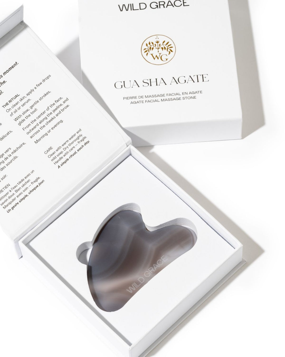 A gua sha facial massage stone in a dark gray translucent finish, presented in an open white gift box with the brand Wild Grace visible on the packaging.
