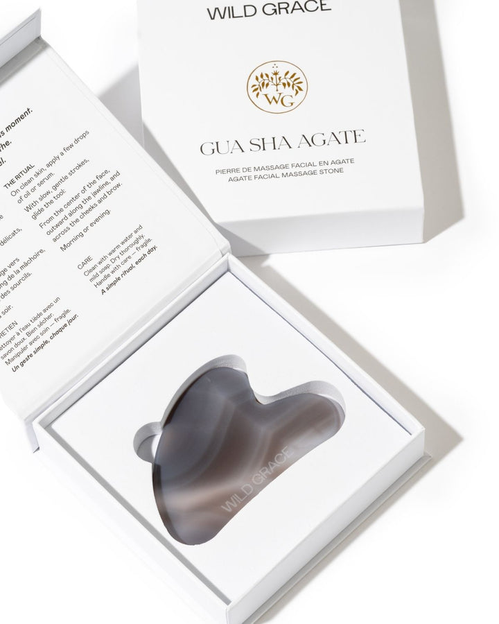A gua sha facial massage stone in a dark gray translucent finish, presented in an open white gift box with the brand Wild Grace visible on the packaging.