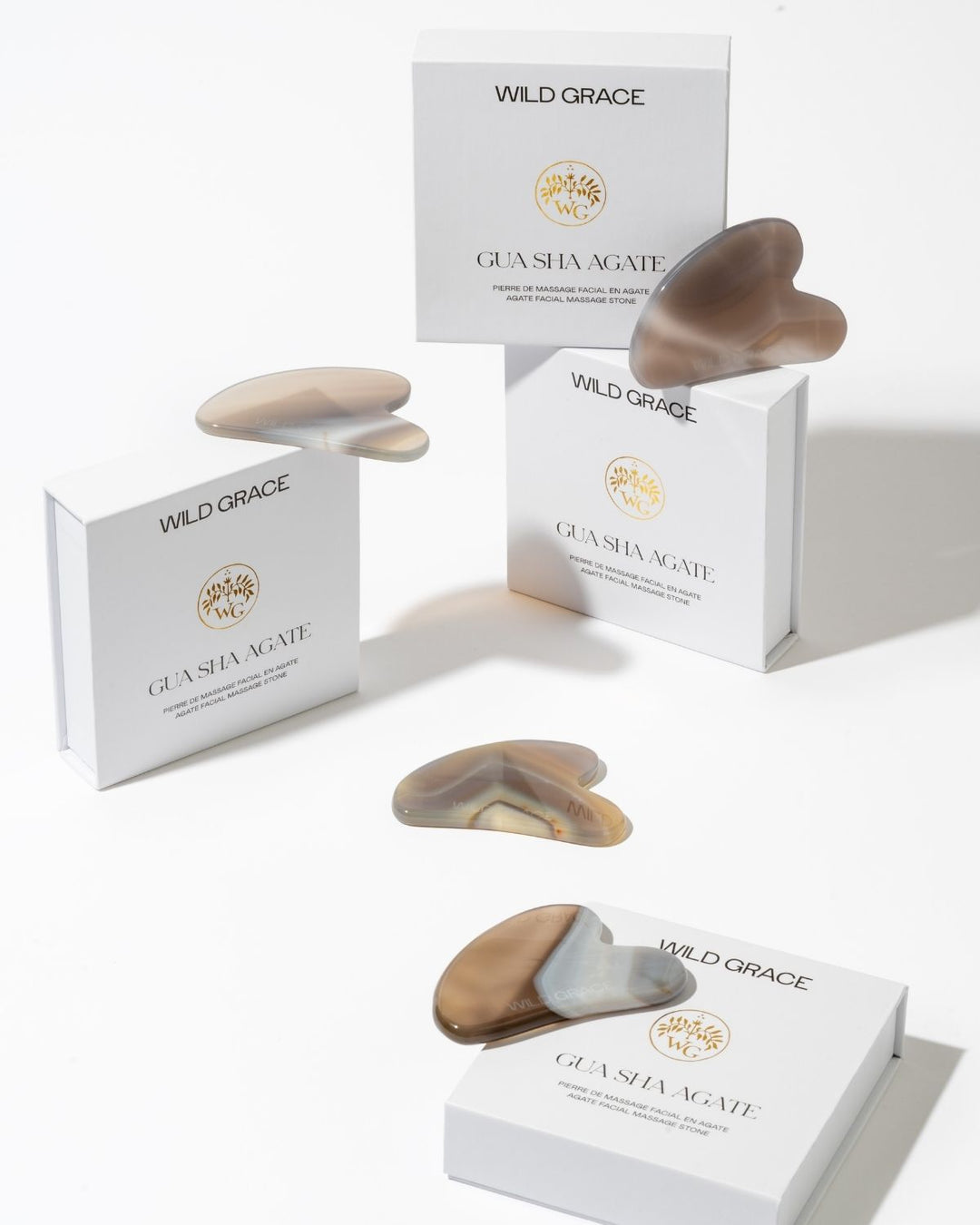 Wild Grace Gua Sha massage stones in various natural stone colors, displayed alongside their white branded packaging boxes on a clean white background.