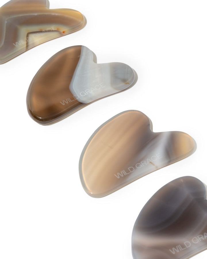 Four gua sha massage tools made from agate with natural banding, arranged diagonally on a white background. The brand name &