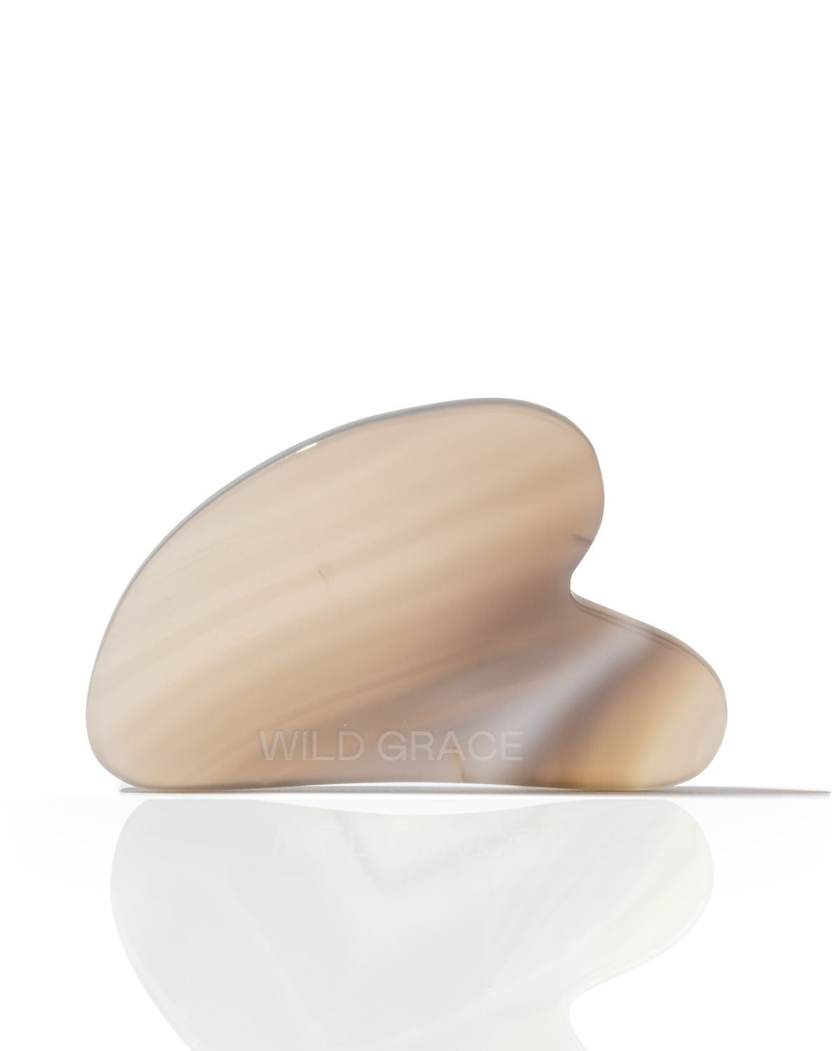 A curved, hand-shaped jade Gua sha massage tool with a translucent beige and light brown marbled pattern, resting on a reflective white surface against a white background. The brand name 'WILD GRACE' is subtly printed on the front.