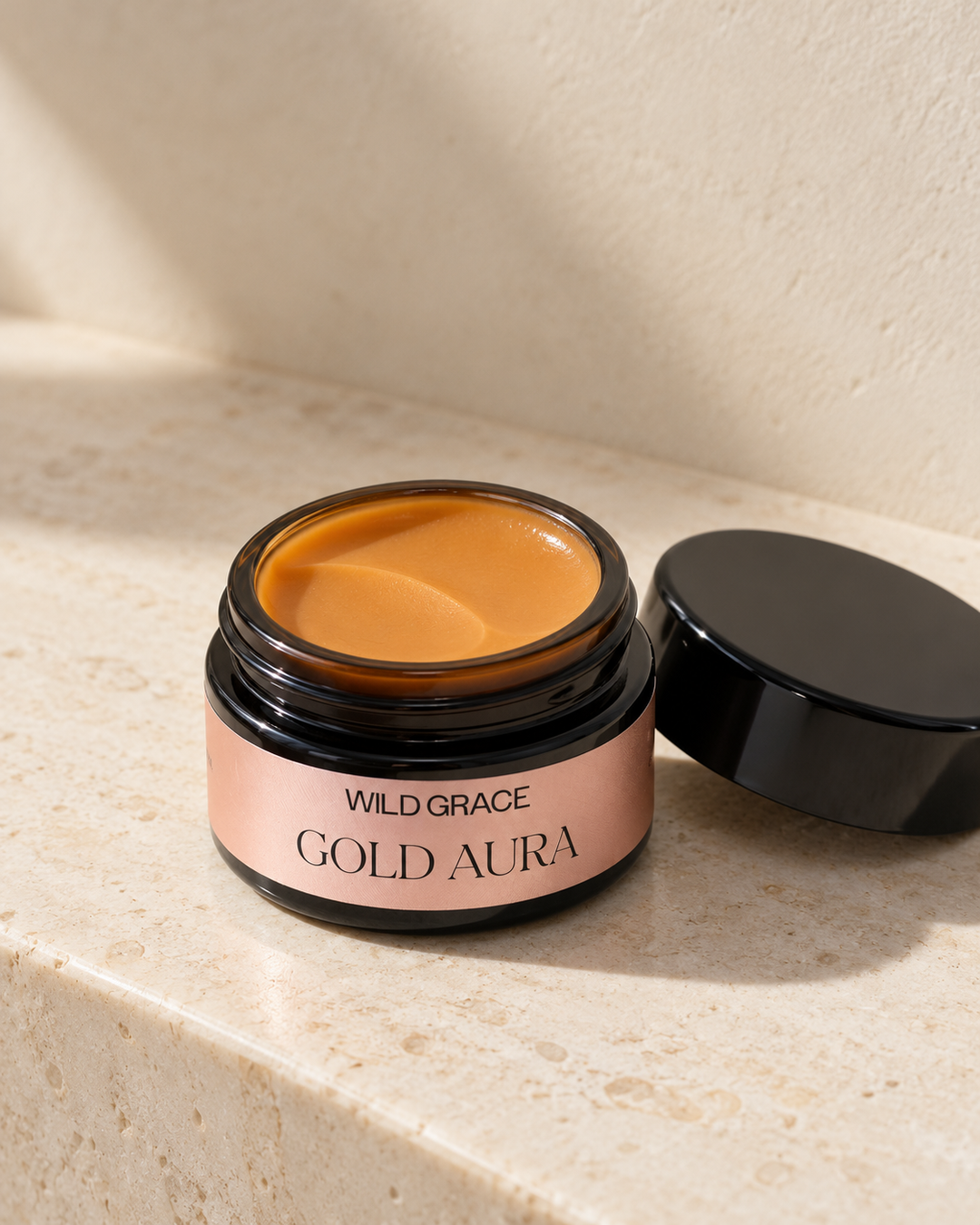 An open cosmetic jar labeled WILD GRACE GOLD AURA sits on a light stone surface with its black lid beside it. The product shows a warm golden-brown cream inside a dark amber jar with a blush pink label.