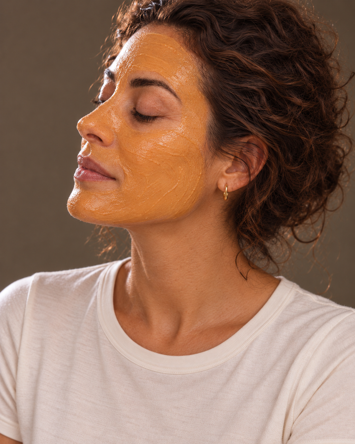A close-up of a woman with a golden-yellow face mask applied, wearing a white crew-neck t-shirt against a solid taupe background.