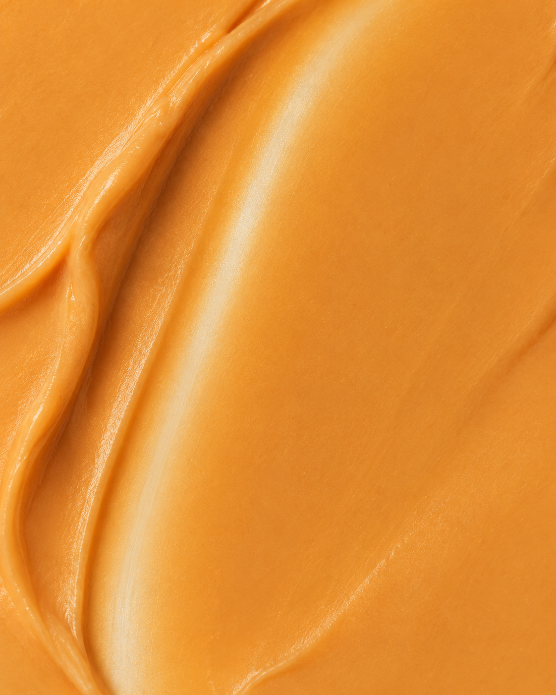 A close-up texture shot of a smooth, creamy cosmetic cream or balm in a warm golden-orange shade, showing soft swirled ridges and a glossy finish.