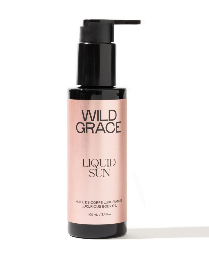A bottle of Wild Grace Liquid Sun Luxurious Body Oil on a white background. The bottle is black with a black pump dispenser and features a light pink label with black text.