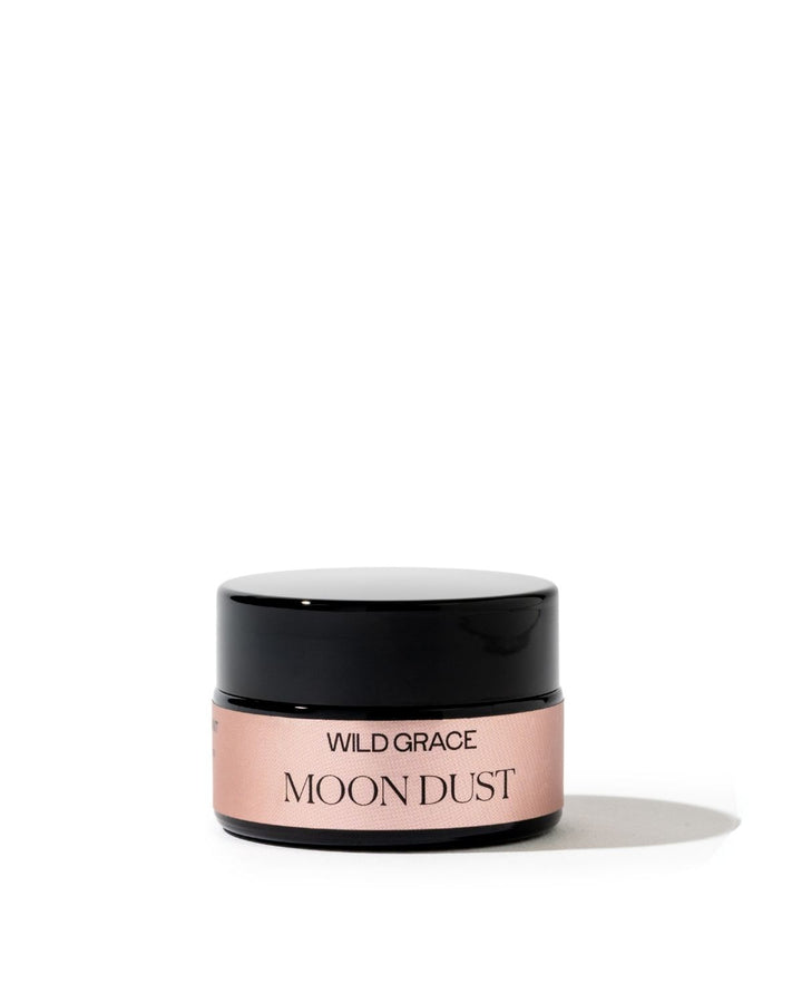 A small cosmetic jar labeled WILD GRACE MOON DUST, featuring a black lid and a dusty pink label, set against a clean white background with a soft shadow.