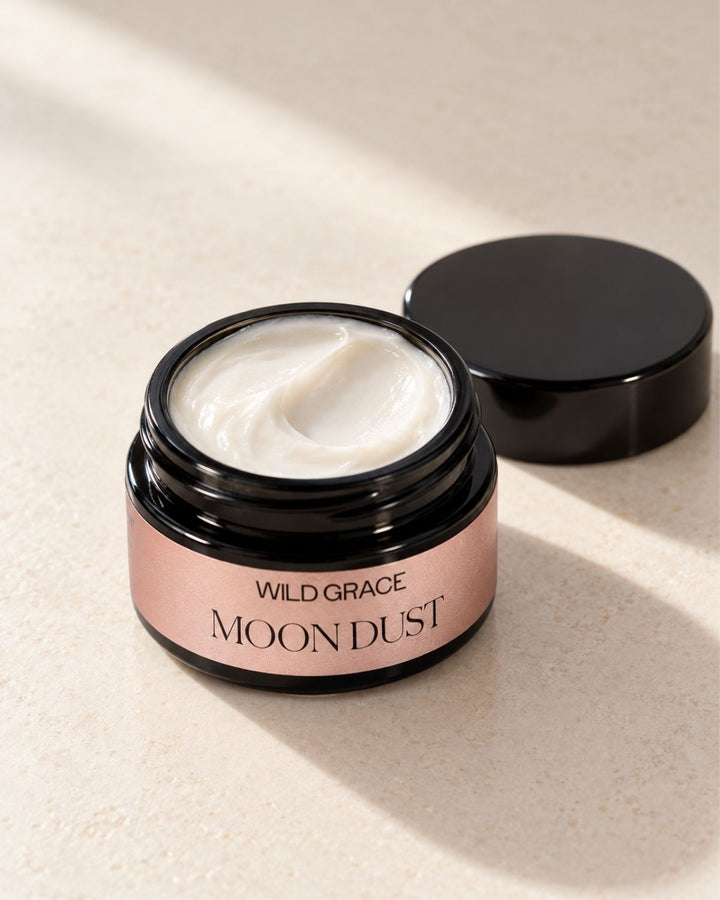 An open jar of Wild Grace Moon Dust cream with its black lid resting beside it on a textured beige surface, featuring soft diagonal shadows.