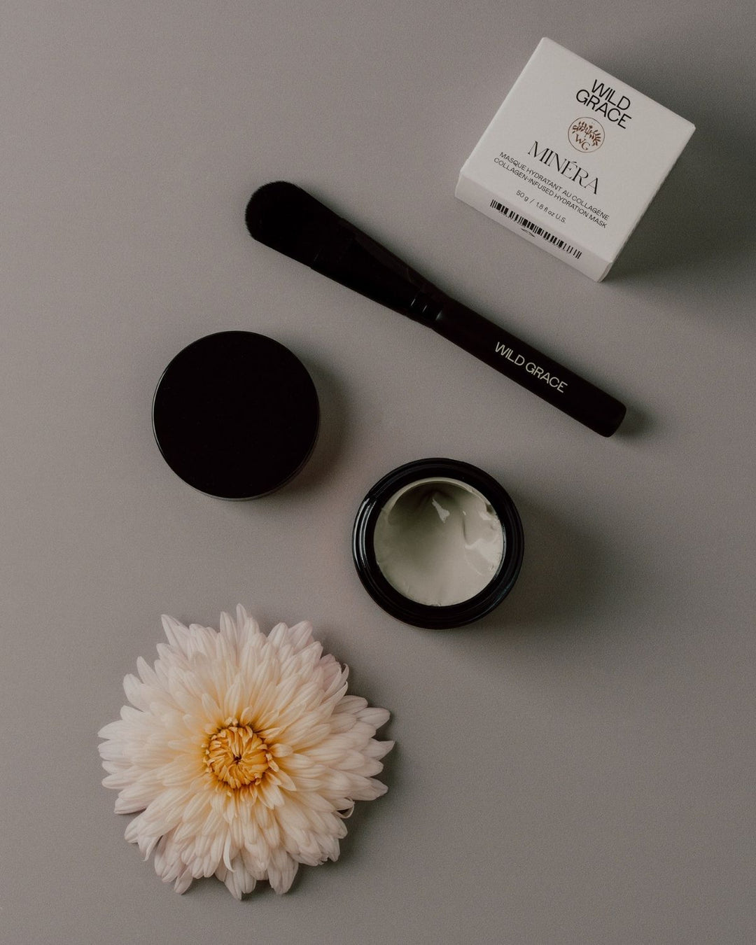 Cosmetic products including a jar, brush, and box on a gray surface with a flower.