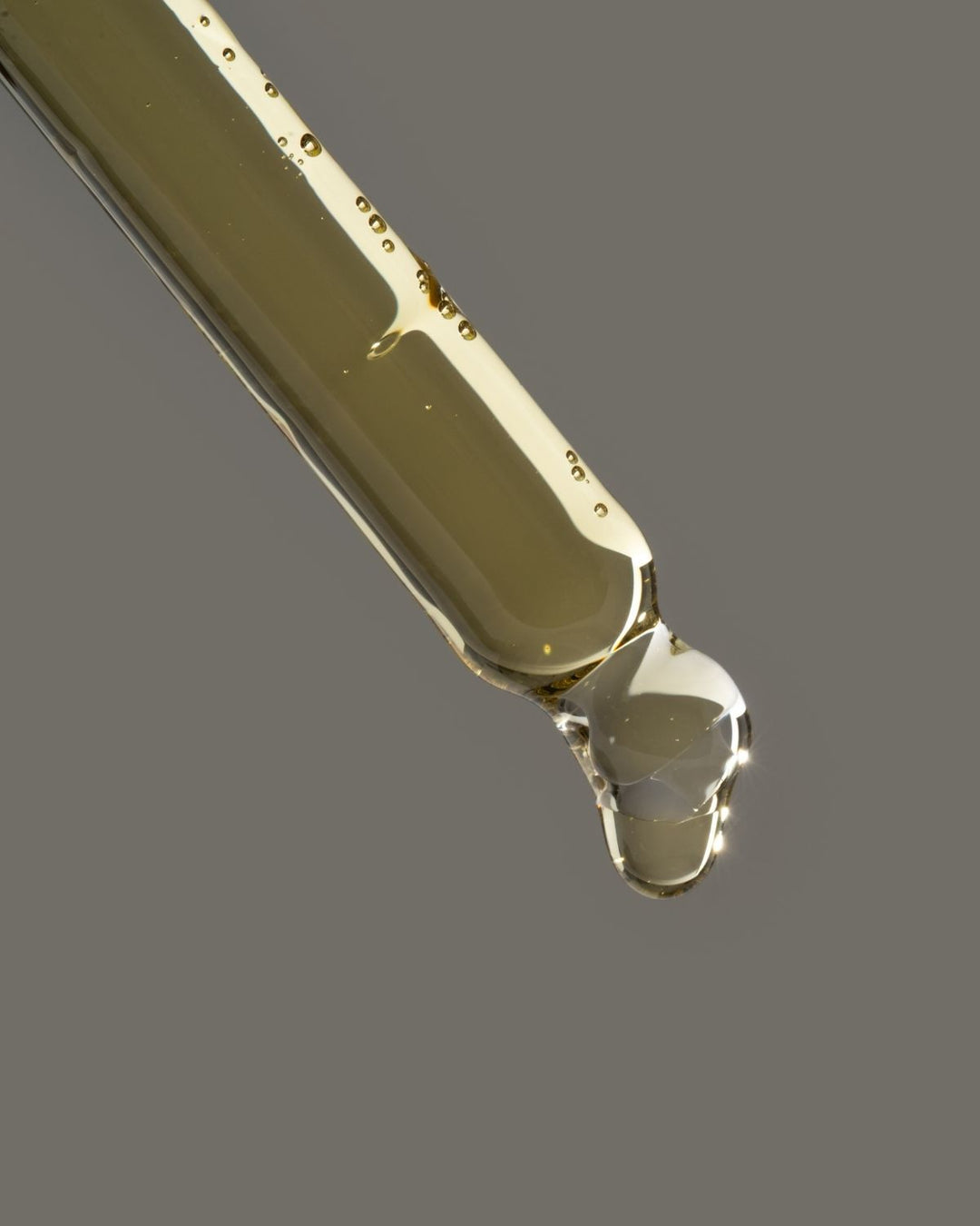 A close-up of a clear glass dropper containing a translucent golden-olive liquid, with a single glossy drop hanging at the tip against a solid gray background.
