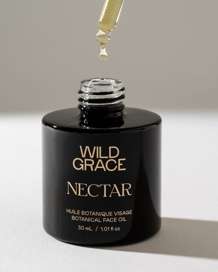 A black bottle of Wild Grace Nectar Botanical Face Oil with gold lettering, shown open with a glass dropper above the bottle, on a neutral light gray background.