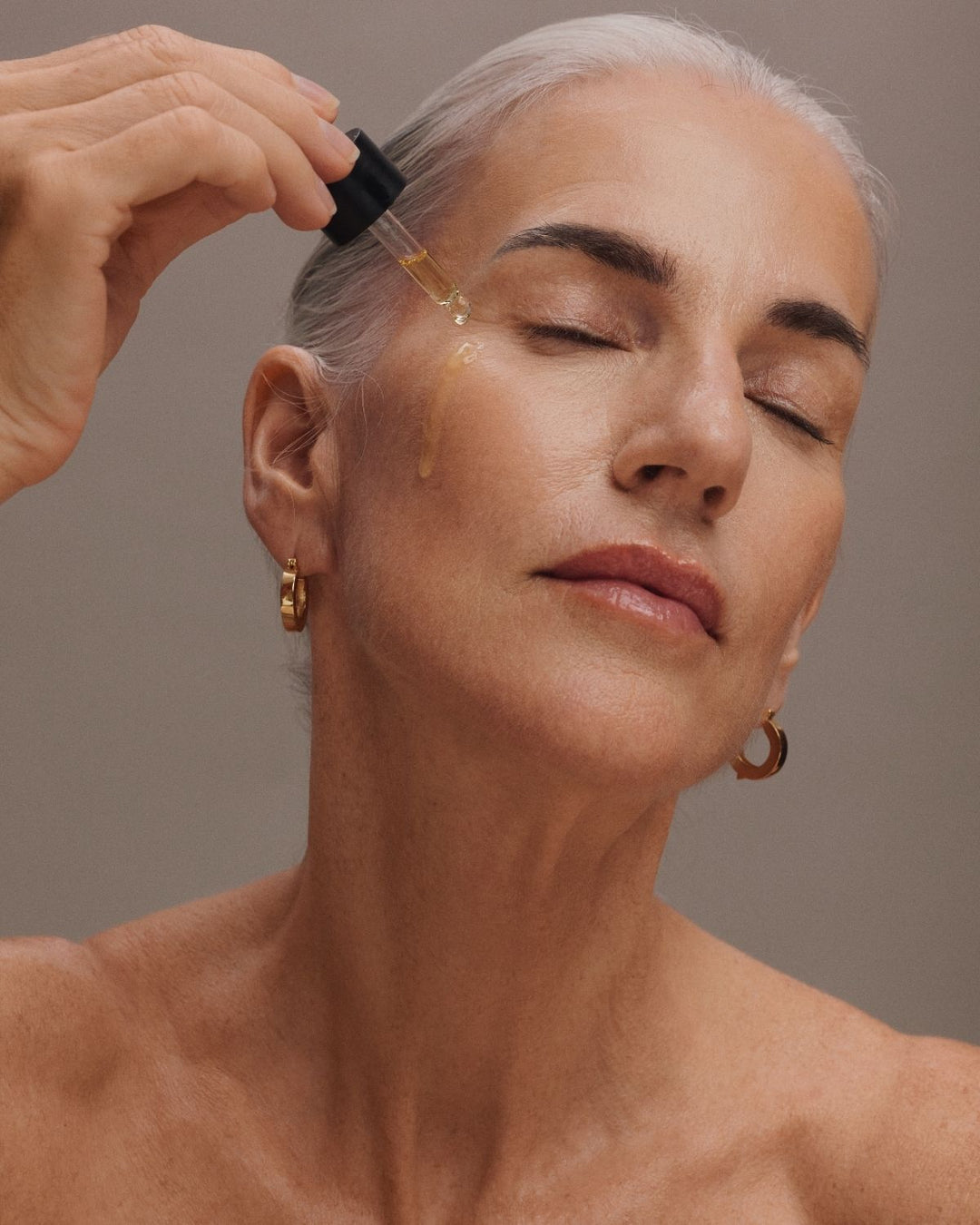 A close-up of a woman with silver hair receiving a dropper treatment, with a hand holding the dropper above her temple. She is wearing small gold hoop earrings against a neutral gray background.