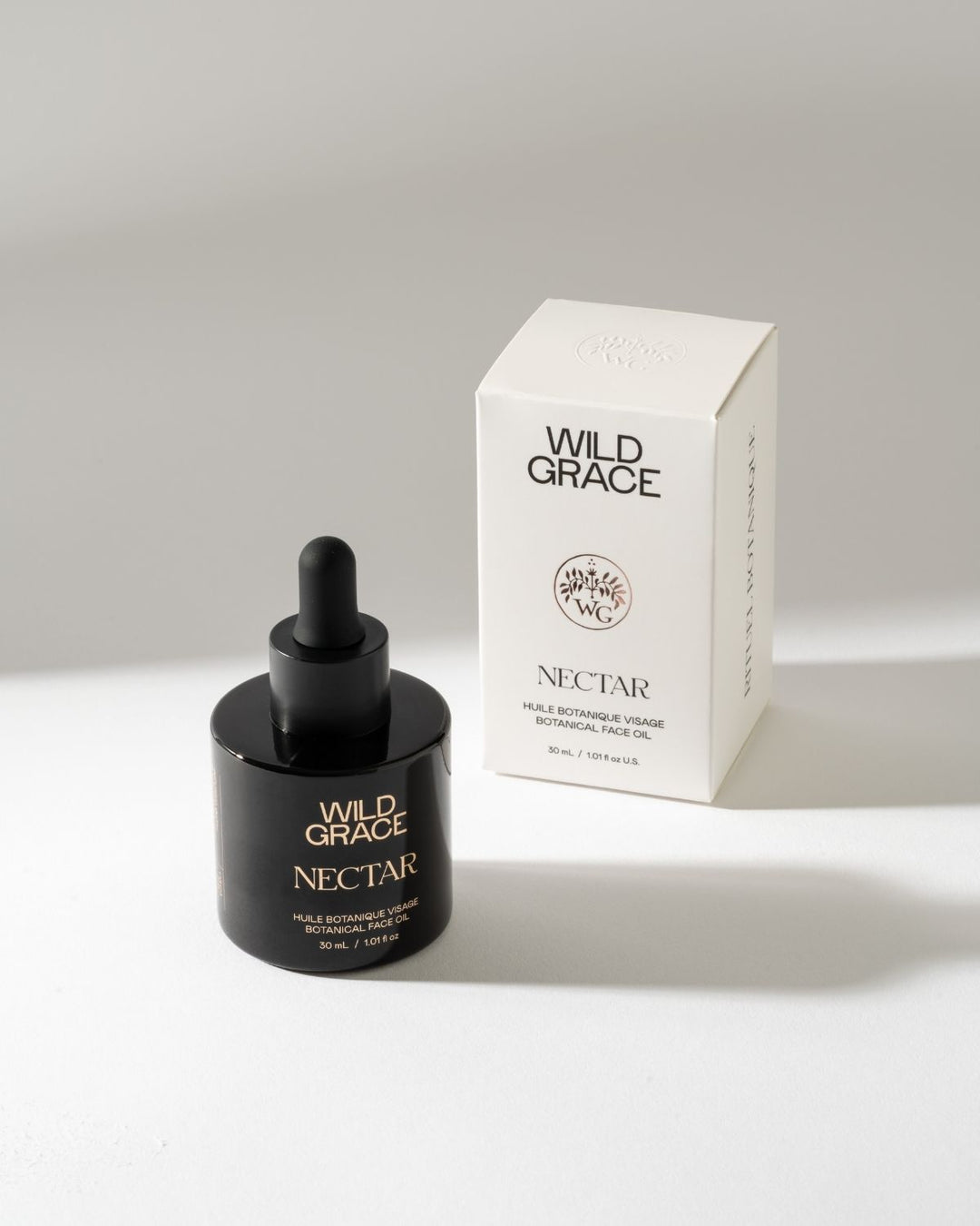 Wild Grace Nectar Botanical Face Oil in a black dropper bottle next to its white packaging box, set against a clean light gray background.