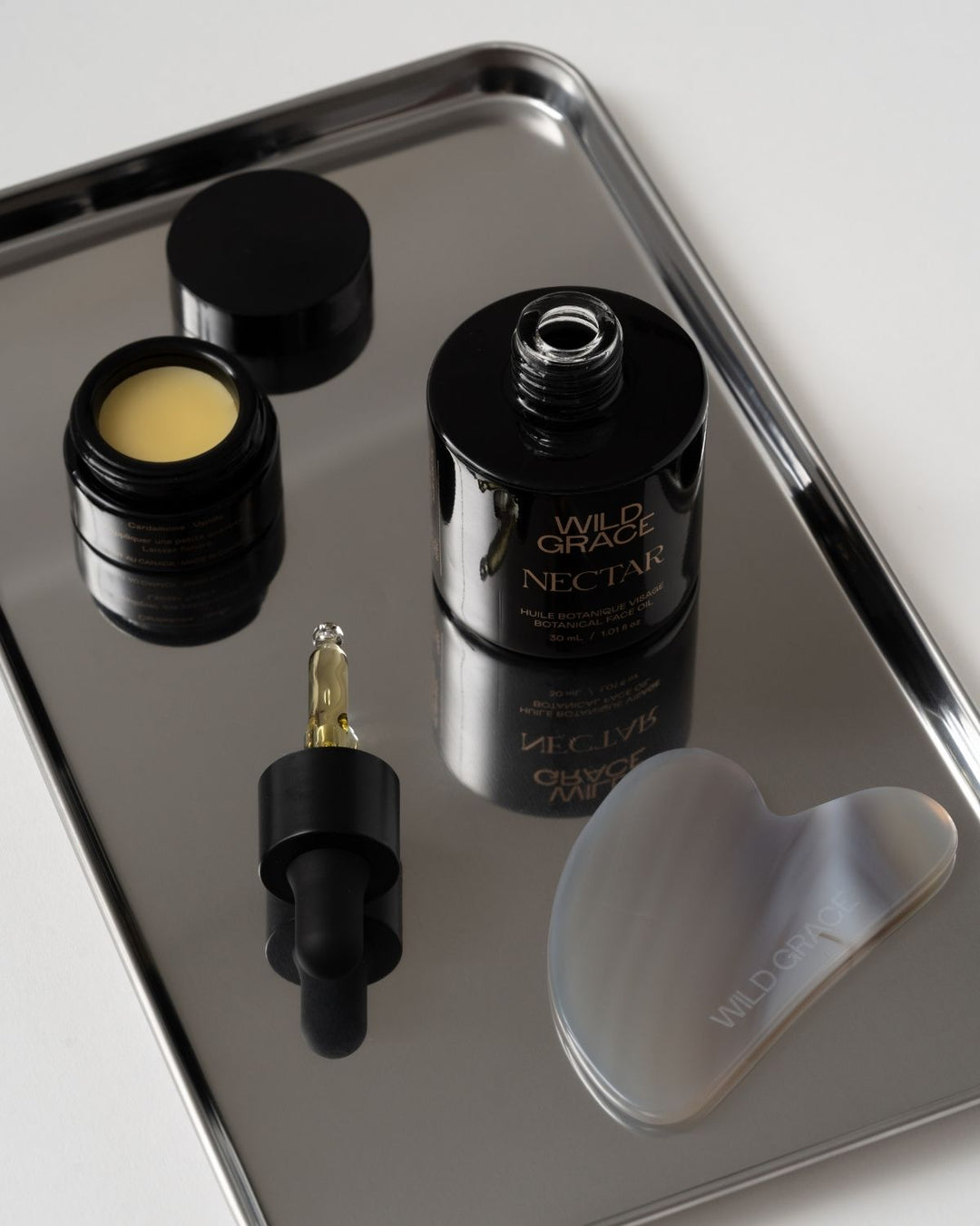 A set of Wild Grace skincare products arranged on a reflective silver tray, including a black bottle labeled Nectar, a small open jar with pale yellow cream, a dropper bottle, and a translucent gray silicone mask.