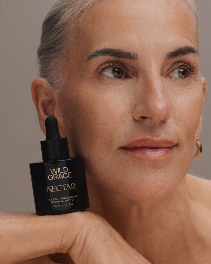 A close-up portrait of a woman holding a black dropper bottle labeled WILD GRACE NECTAR against a soft taupe background.