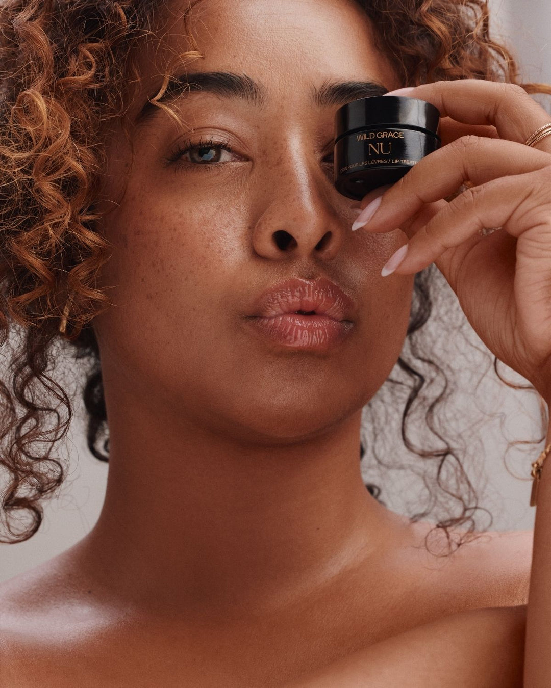 A close-up lifestyle shot of a woman with curly hair holding a small black jar of Wild Grace NU Retinol Serum against her eye, showcasing the product in use.