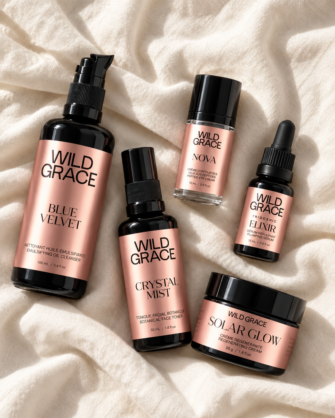 Five Wild Grace haircare products arranged on a cream-colored textured fabric background. The products include a Blue Velvet shampoo bottle, a Crystal Mist spray bottle, a Nova conditioner bottle, an Elixir dropper bottle, and a Solar Glow treatment jar, all featuring rose gold labels and black caps.