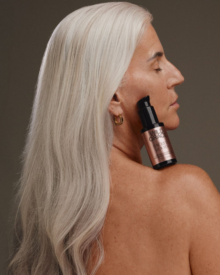A woman with long platinum blonde hair shown in profile, holding a small bottle of Morphe Hair Growth Serum against her neck, against a dark brown background.