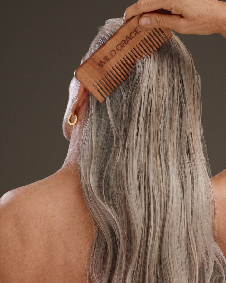 A close-up lifestyle shot of a woman with long, wavy grayish-blonde hair being combed from the back with a wooden comb engraved with the brand name &