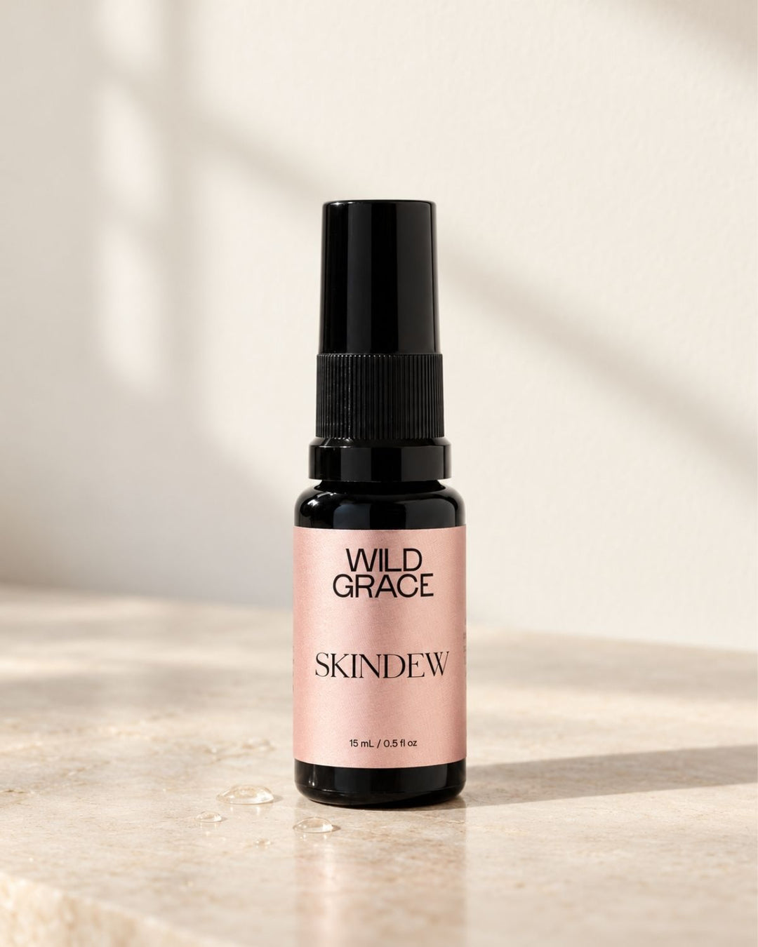 A small bottle of Wild Grace Skindew spray with a black pump and a dusty pink label, standing on a light stone surface with soft window shadows.