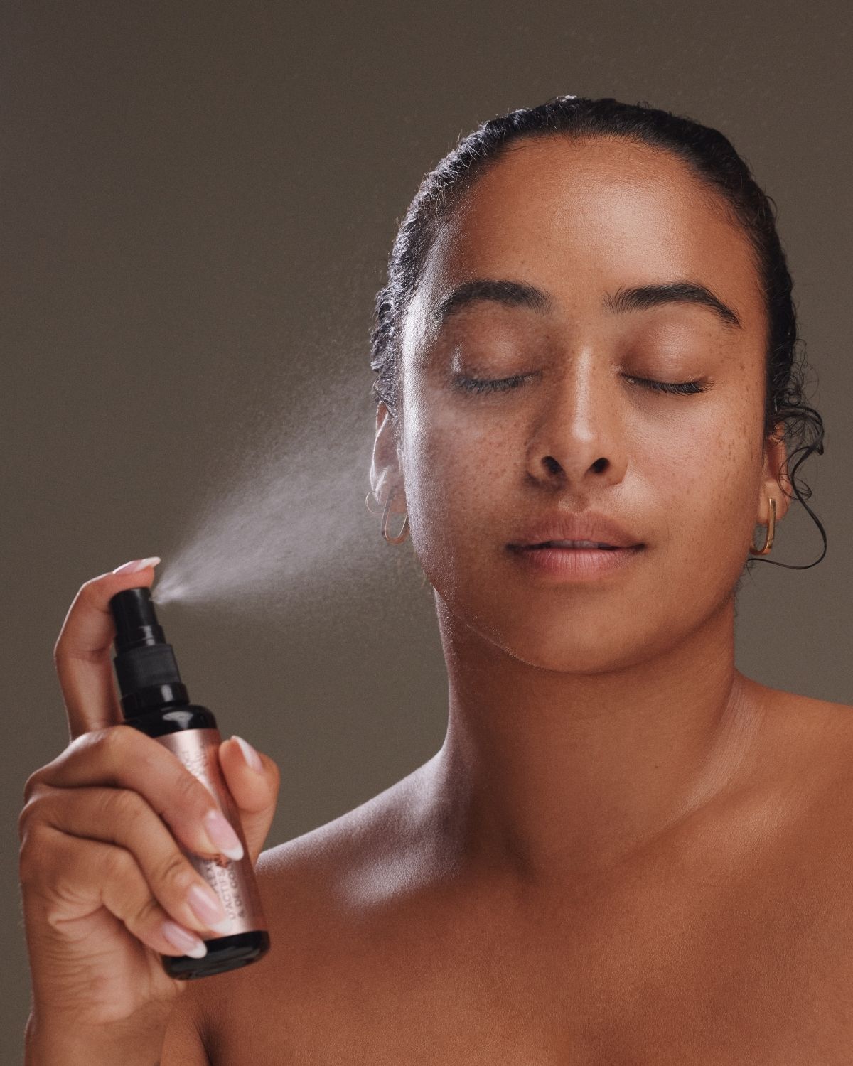 A woman with her eyes closed holds a small spray bottle in front of her face, releasing a fine mist. She is shown from the shoulders up against a plain taupe-brown background.