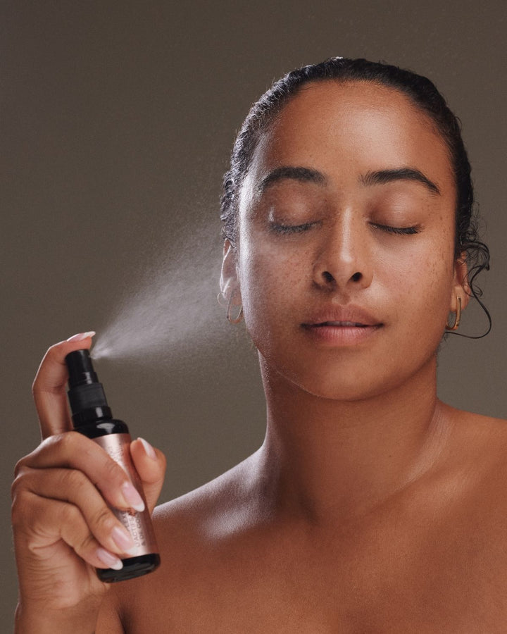 A woman with her eyes closed holds a small spray bottle in front of her face, releasing a fine mist. She is shown from the shoulders up against a plain taupe-brown background.