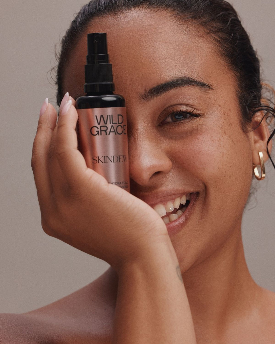 A smiling woman holds a bottle of Skincare Wild Grace spray next to her face against a neutral beige background.