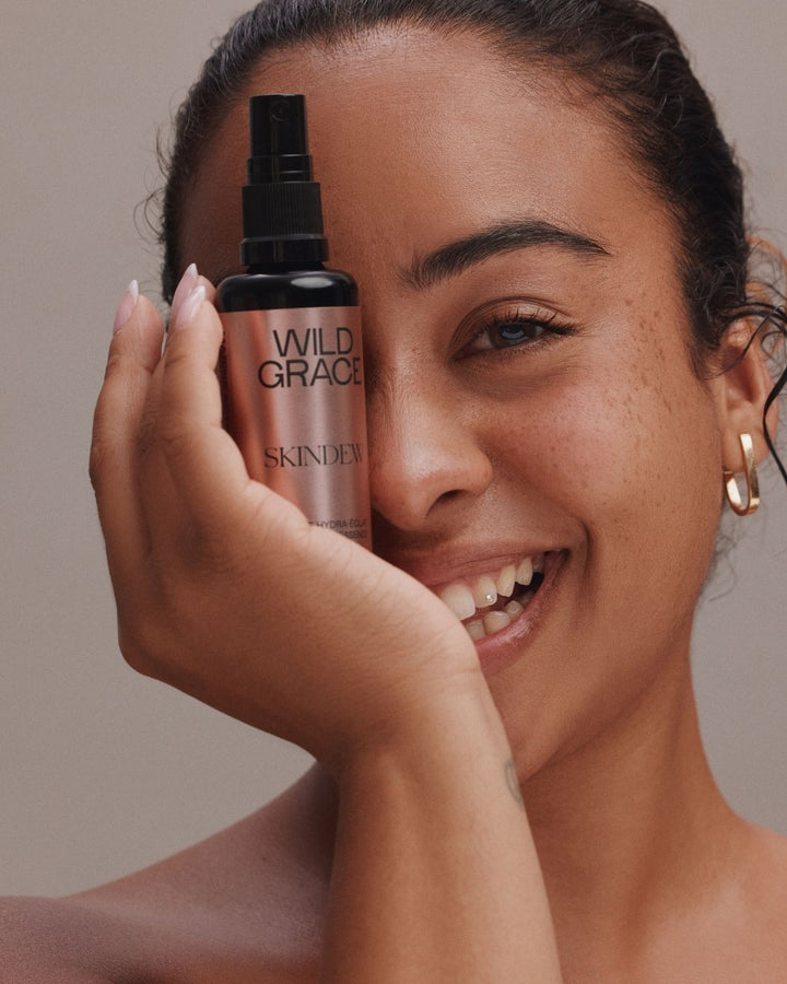 A smiling woman holds a bottle of Skincare Wild Grace spray next to her face against a neutral beige background.