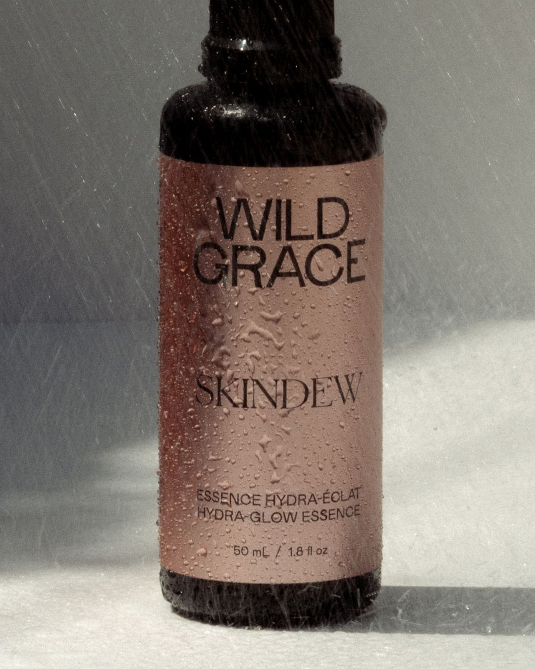 A dark amber bottle of Wild Grace Skinew Hydra-Glow Essence covered in water droplets, set against a gray background with a soft light reflection.