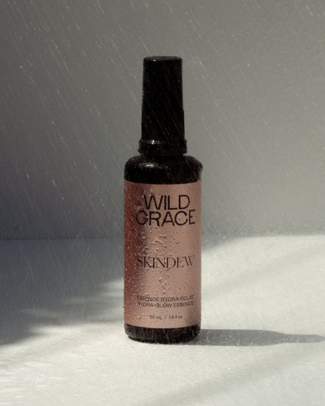 A bottle of Wild Grace Skindew Essence Hydra Color spray, featuring a dark brown glass bottle with a black cap and a dusty rose pink label, set against a gray background with a white textured surface and light rain falling.