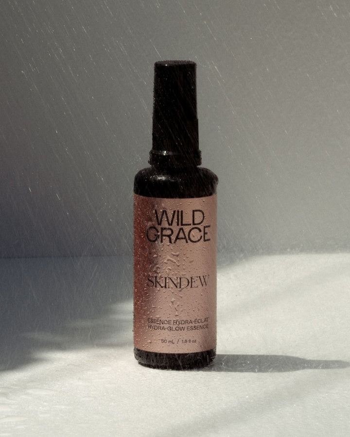 A bottle of Wild Grace Skindew Essence Hydra Color spray, featuring a dark brown glass bottle with a black cap and a dusty rose pink label, set against a gray background with a white textured surface and light rain falling.