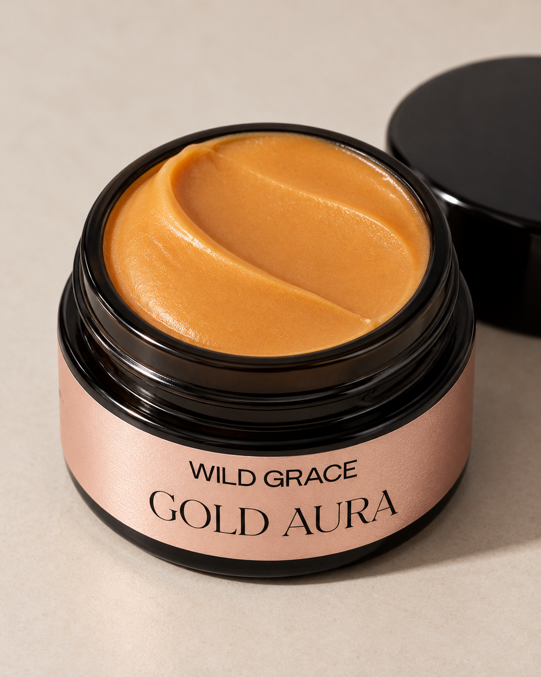 An open jar of Gold Aura Wild Grace skincare cream showing the smooth, golden-brown product inside, with its black lid resting beside it on a neutral beige background.