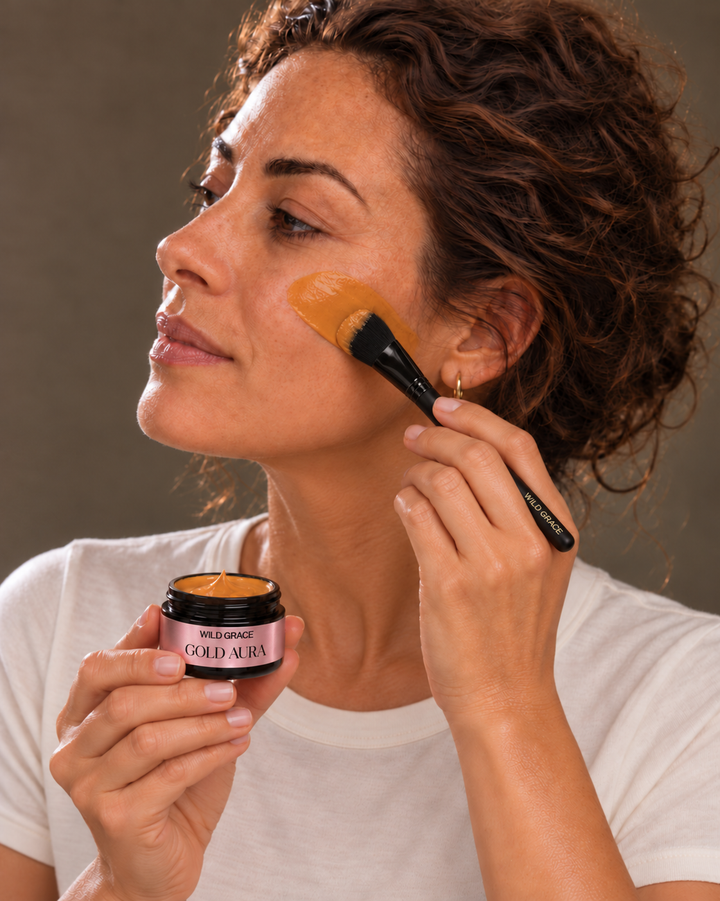 A woman applies a yellow-orange facial mask or cream with a brush while holding an open jar labeled Wildpflanz Good Avenir.