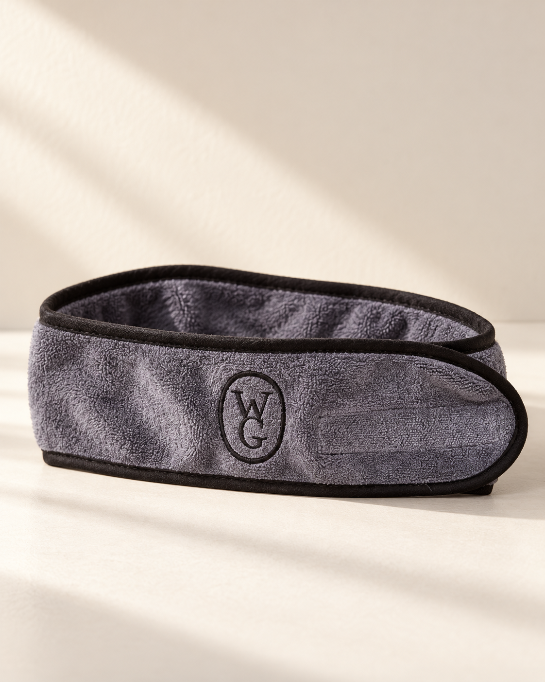 A gray textured microfiber eye mask with black trim and an embroidered circular 'WCG' logo on the front, displayed on a warm beige surface with soft diagonal shadows.