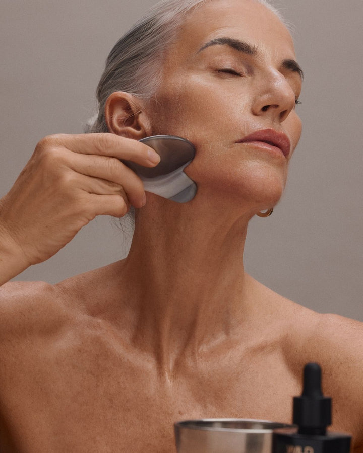 A close-up lifestyle shot of a woman with gray hair applying a skincare product using a gray silicone device on her neck and jawline. A small dropper bottle is visible in the foreground against a neutral gray background.