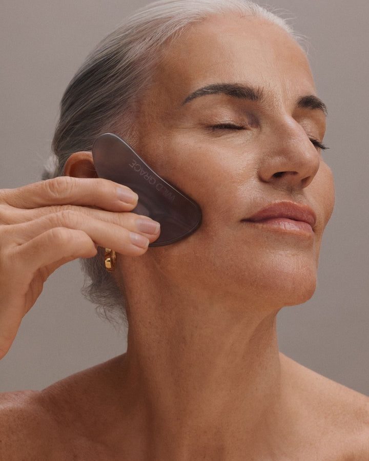 A close-up of a woman with gray hair using a dark gray facial gua sha massage tool on her cheek, set against a neutral gray background.