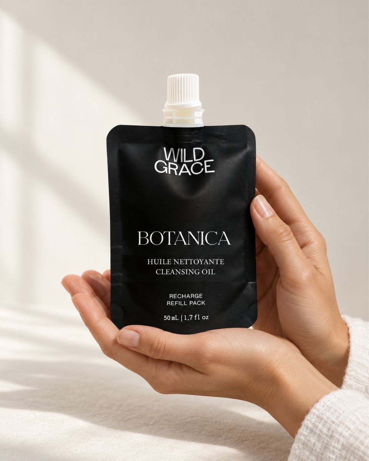 A black refill pouch of Wild Grace Botanica Cleansing Oil with a white cap, set against a warm beige background with soft window shadows.