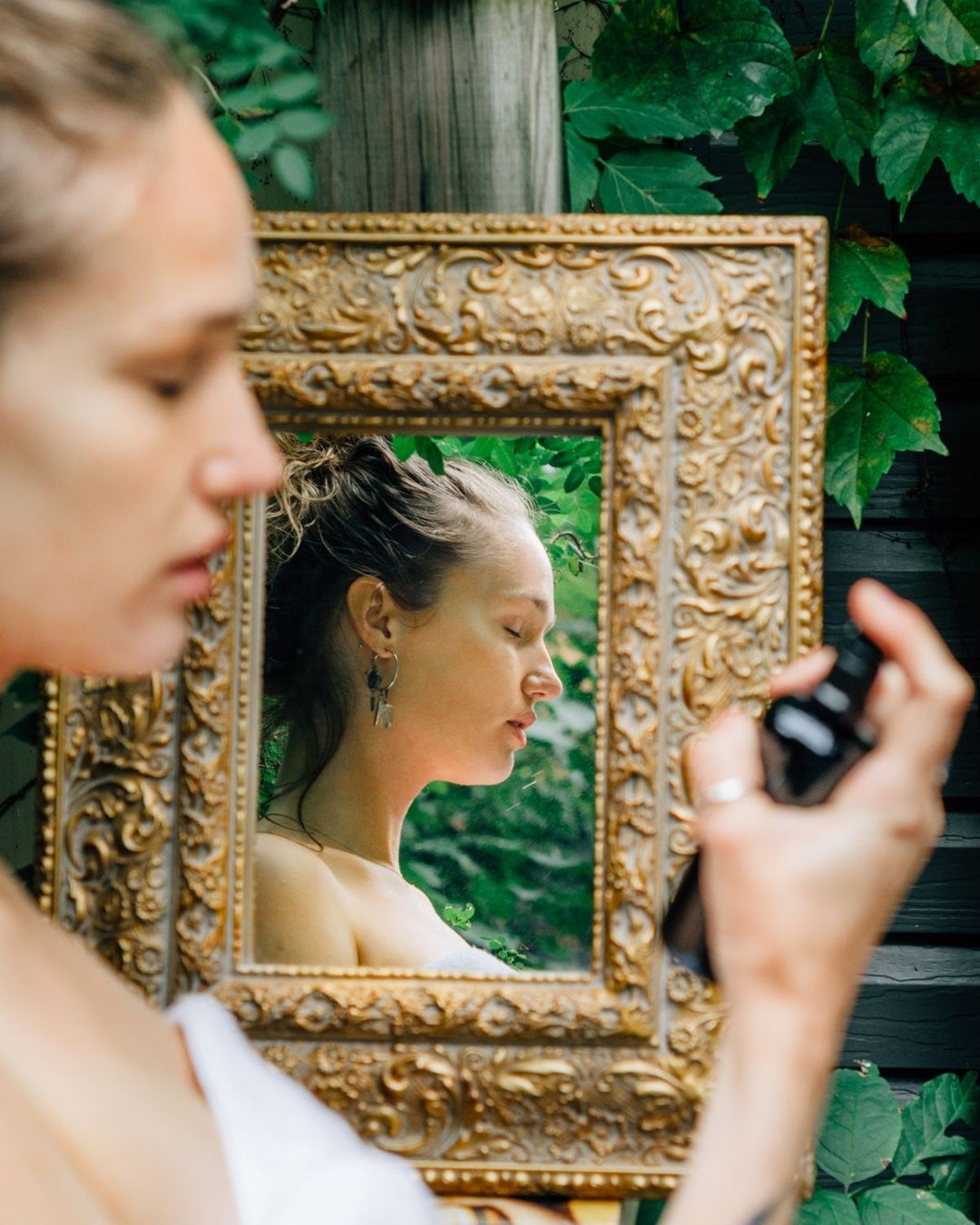 Woman misting her face with the crystal mist toner with ornate gold mirror surrounded by green foliage.