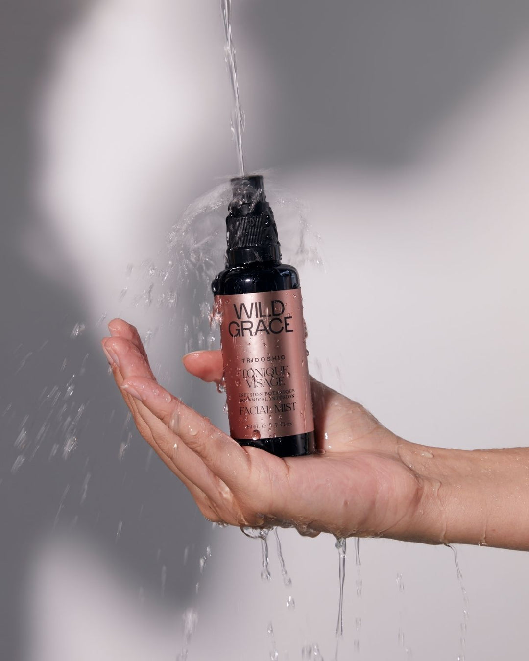 A hand holding a bottle of Wild Grace Hydration Primer under a stream of water, with water droplets splashing around the bottle against a gray background.