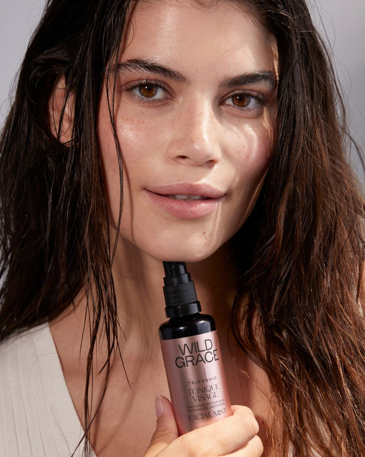 A close-up of a woman with wet, styled dark hair holding a bottle of Wild Grace Haircare Hair Reviver &amp; Volume Treatment against a neutral gray background.