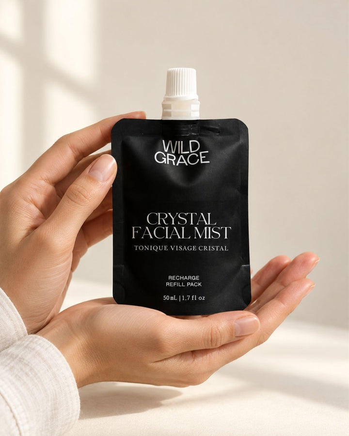 A person holds a black refill pouch of Wild Grace Crystal Facial Mist against a warm beige background with soft sunlight and shadows.