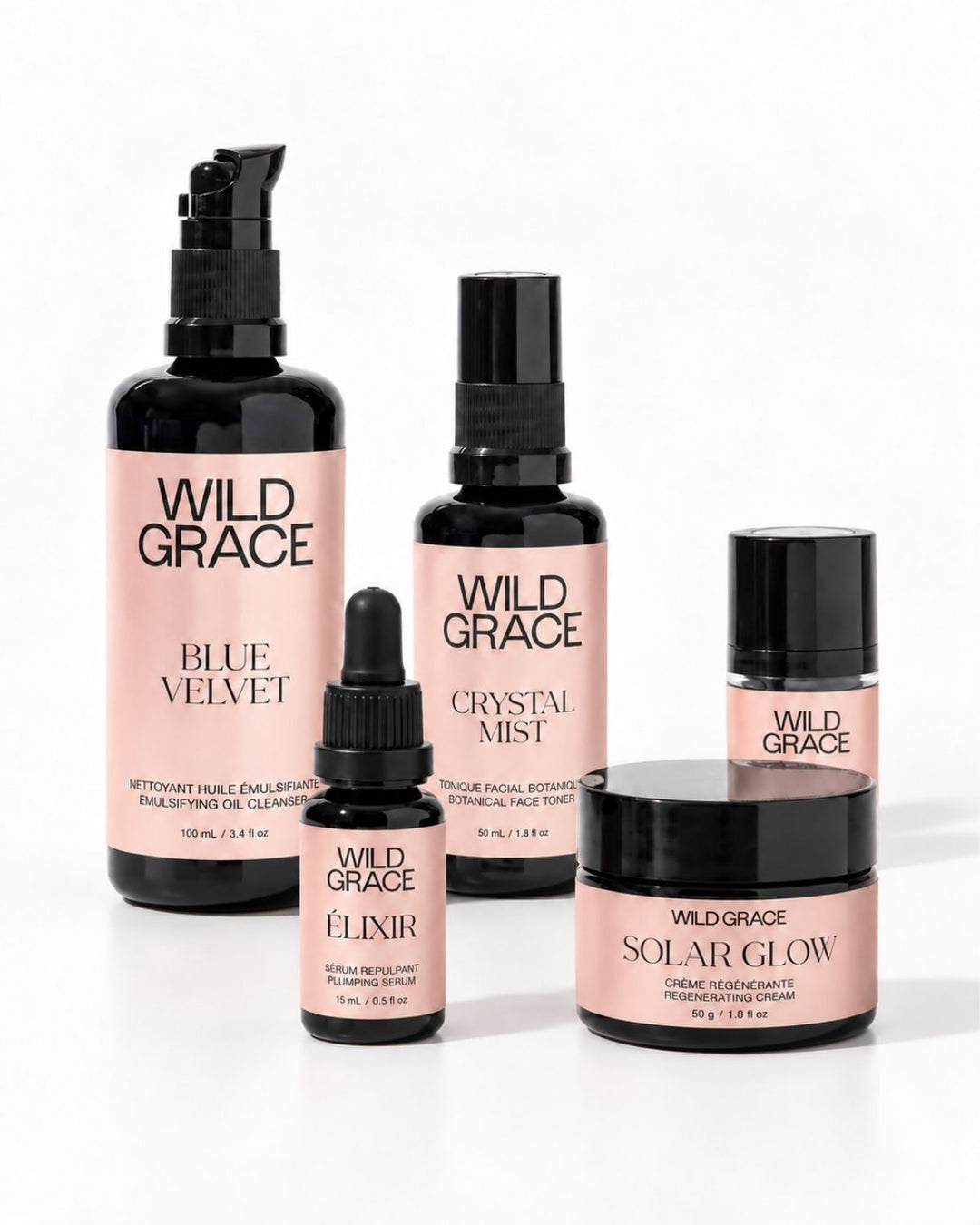A set of five Wild Grace skincare bottles and jars arranged on a white background, featuring black packaging with soft pink labels. The products include Blue Velvet, Crystal Mist, Nova, Elixir, and Solar Glow.